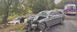 Miejsce wypadku drogowego. Uszkodzony biały samochód znajduje się poza jezdnią pod drzewem, a na drodze stoi samochód z oznaczeniem TAXI. Na jezdni i poboczu widoczne są elementy uszkodzonych pojazdów. W tle na drodze widać karetkę pogotowia ratunkowego.