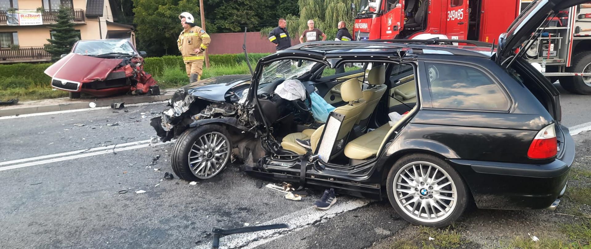 Dwa samochody osobowe uszkodzone stoją na drodze pod skosem po przeciwnych stronach drogi. W tle widoczny samochód pożarniczy oraz strażacy.