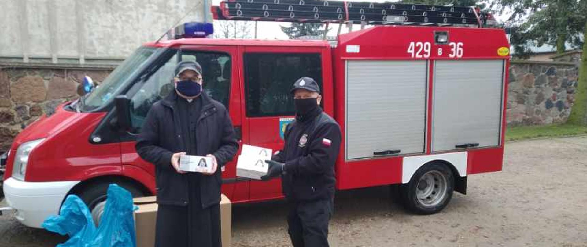 Strażak OSP oraz ksiądz. W tle samochód pożarniczy.