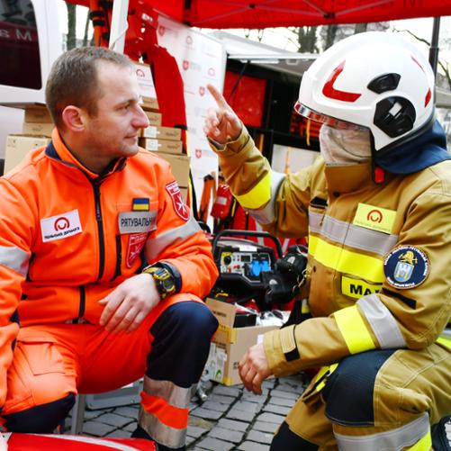 Strażak i ratownik medyczny kucają przy samochodzie strażackim.