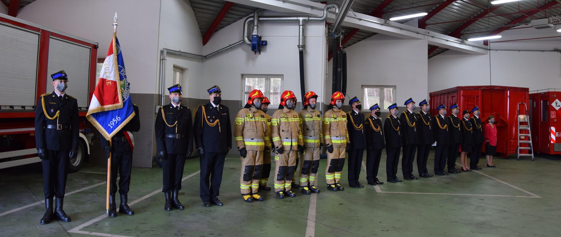 W szeregu, podczas apelu, który odbywa się w garażu, stoją strażacy - poczet sztandarowy, dowodca, załoga w umundurowaniu służbowym, następnie załoga w umundurowaniu galowym. W tle pojazdy i sprzet pożarniczy.