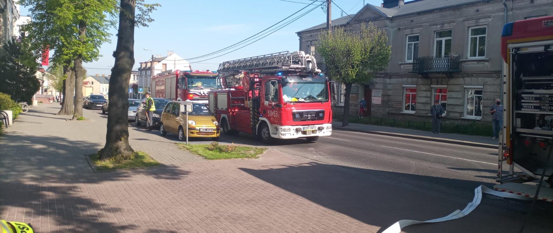 Zdjęcie przedstawia 3 samochody pożarnicze. Znajdują się na ulicy, w tle widać inne pojazdy osobowe oraz budynki mieszkalne. Z jednego samochodu strażackiego rozwinięta jest linia wężowa oraz szybkie natarcie. 