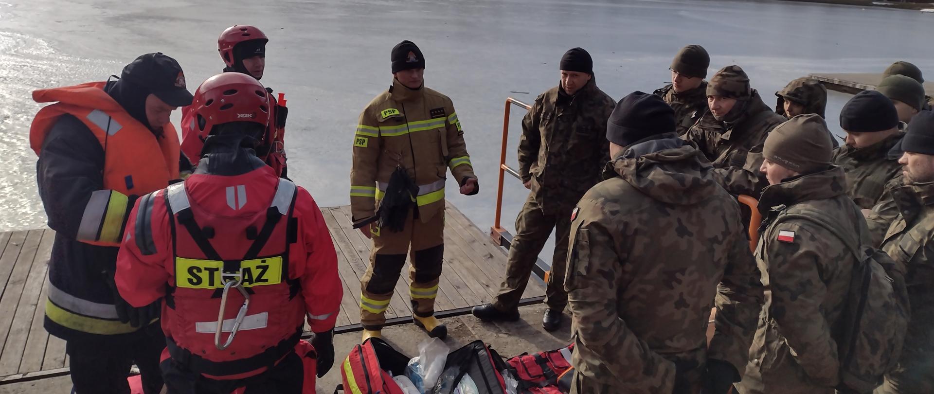 Na zdjęciu widać na pomoście zbiornika wodnego strażaków oraz żołnierzy WOT. Strażacy pokazują sprzęt i wyjaśniają zasady działań ratownictwa lodowego. Trzech strażaków ubranych jest w sprzęt do podejmowania osób z wody.