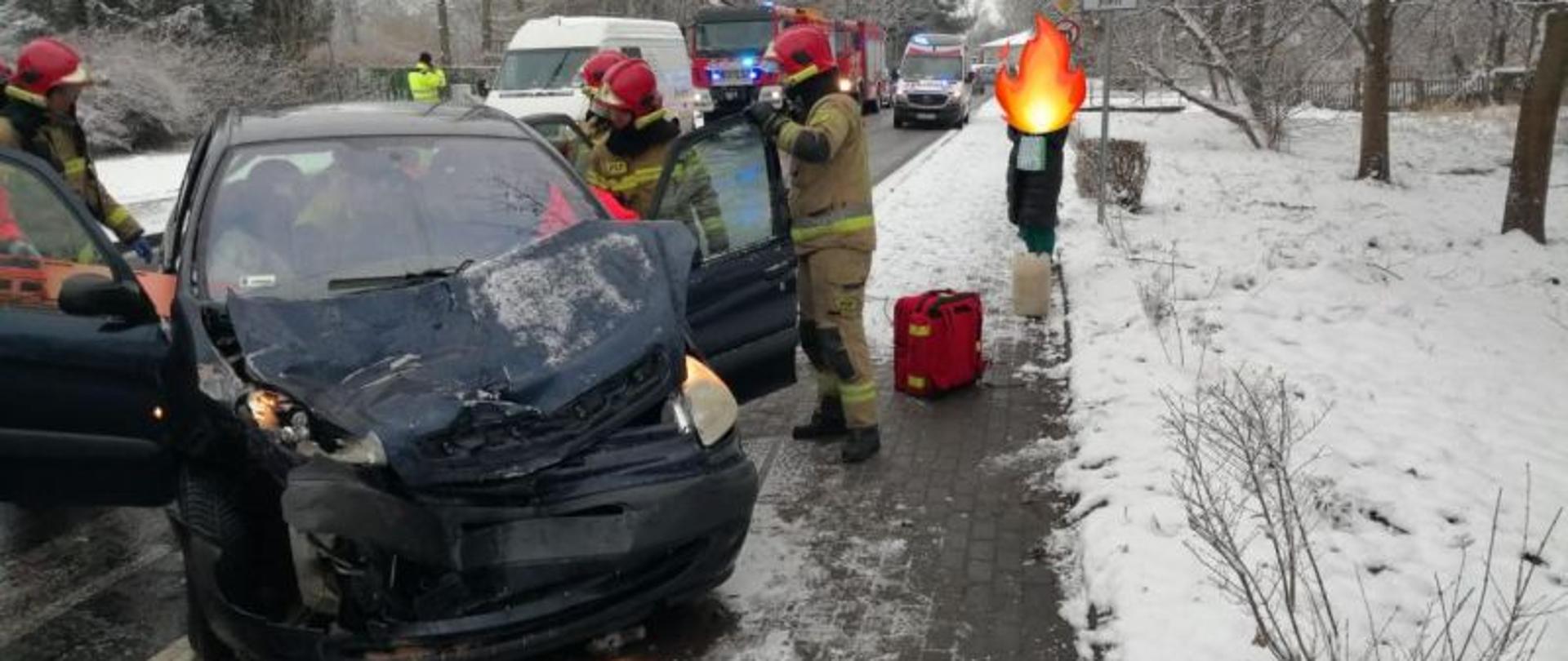 Widok na rozbite auto osobowe. Sceneria zimowa, auto ma mocno zniszczony przód. W tle pojazdy stojące w małym korku. Przy aucie strażacy.
