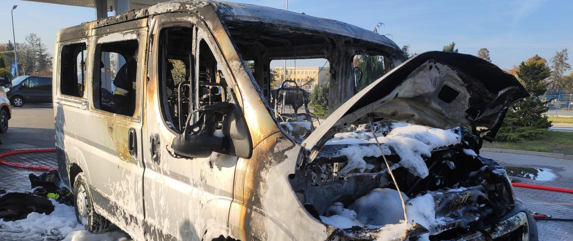 Biały samochód typu bus stoi na placu koło stacji paliw z otwarta pokrywą silnika wokół niego widoczna piana gaśnicza