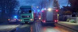 Po lewej jest radiowóz Policji, na środku drogi stoi zielony samochód ciężarowy, po prawej samochody straży pożarnej, jest ciemno