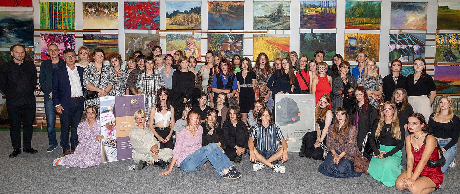 Zdjęcie grupowe przedstawiające uczniów oraz wychowawców na wernisażu pleneru Rychwał XXIX na tle rozwieszonych obrazów namalowanych na plenerze. 