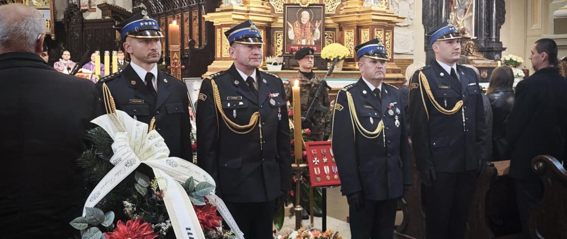 Strażacy w mundurach galowych stoją na baczność przed trumną śp. płk. poż. Stanisława Luli. Obok widoczny wieniec z biało-czarną wstęgą pożegnalną oraz medalami i odznaczeniami zasłużonego oficera.
