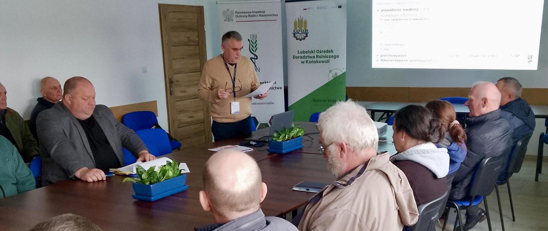 Grupa osób siedzi przy stole konferencyjnym. Jeden mężczyzna stojąc czyta z kartki trzymanej w ręku. W tle na ścianie wyświetlana jest prezentacja