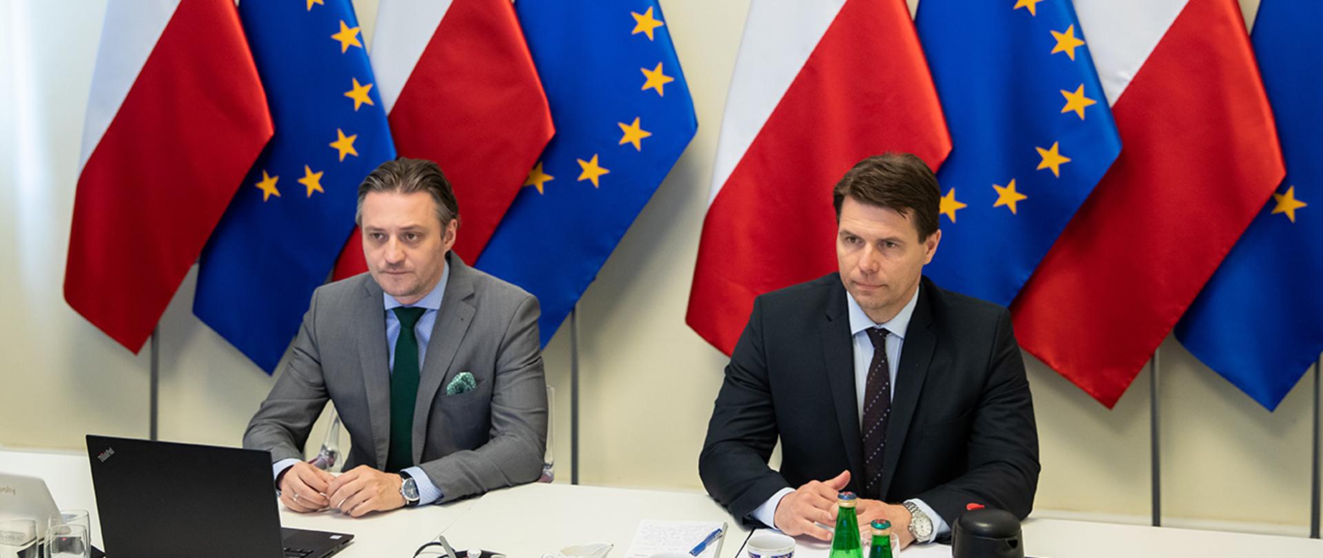 Na zdjęciu widać siedzących przy stole Bartosza Grodeckiego wiceministra SWiA i Mariusza Boguszewskiego dyrektora Departamentu Spraw Międzynarodowych MSWiA. W tle widać flagi Polski i UE.