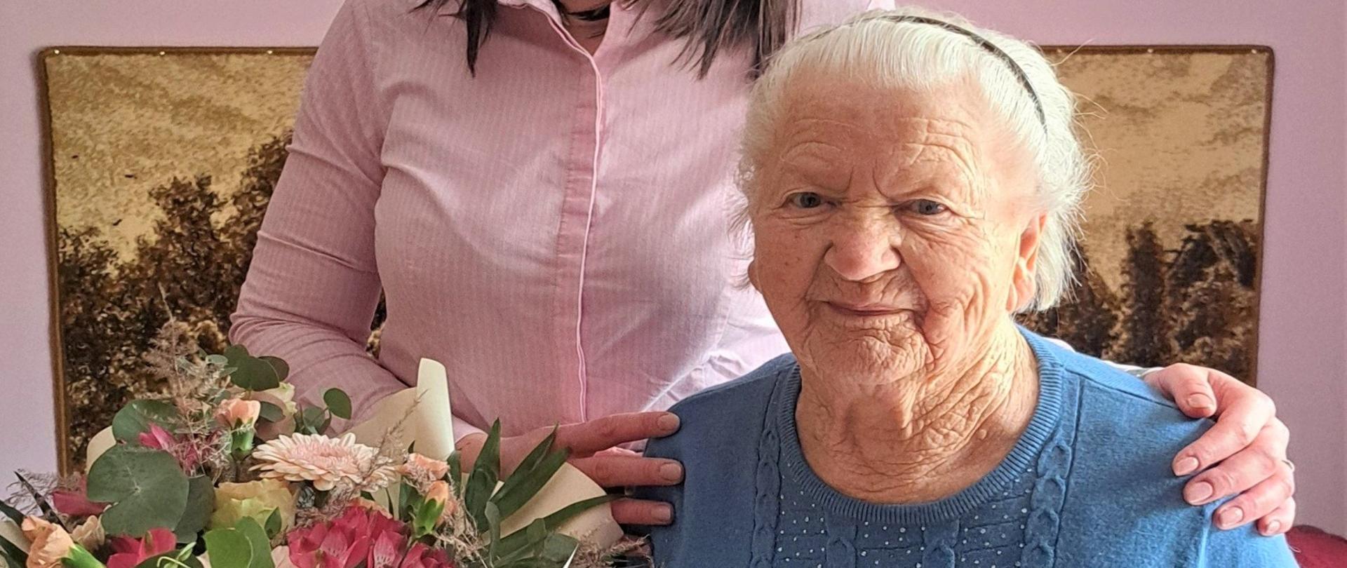 Młoda kobieta w różowej koszuli stoi za starszą kobietą z siwymi włosami i trzyma rękę na jej ramieniu. Pani Władysława trzyma w rękach bukiet kwiatów. 