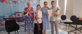 Na zdjęciu cztery osoby pozują na tle dekoracji w morskim stylu przygotowanej z okazji uroczystości. W centrum stoi młoda skrzypaczka trzymająca skrzypce i smyczek oraz dyplom z podziękowaniem. Obok znajdują się trzy osoby dorosłe – dwie z nich trzymają pamiątkowe dyplomy i upominki. Po lewej stronie widoczny jest bukiet kwiatów. Po prawej stronie ustawiono instrument klawiszowy przygotowany do akompaniamentu. Nad uczestnikami znajdują się niebiesko-białe balony i dekoracje nawiązujące do wakacyjnego, nadmorskiego klimatu. Zdjęcie oddaje uroczystą i serdeczną atmosferę wydarzenia.