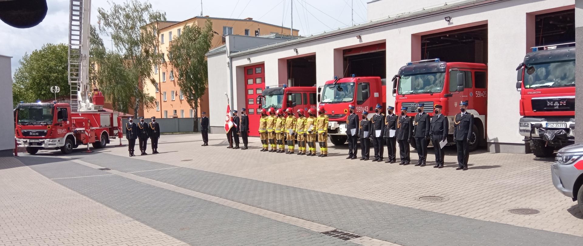 Po lewej jest podnośnik straży pożarnej, przed nim jest trzech strażaków z pocztu flagowego, na środku jest sztandar i strażacy podczas uroczystego apelu, za nimi cztery czerwone samochody straży 