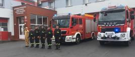 Zdjęcie przedstawia strażaków którzy stoją w szeregu. Jeden z nich oddaje honor poprzez salutowanie. W tle stoją dwa wozy strażackie