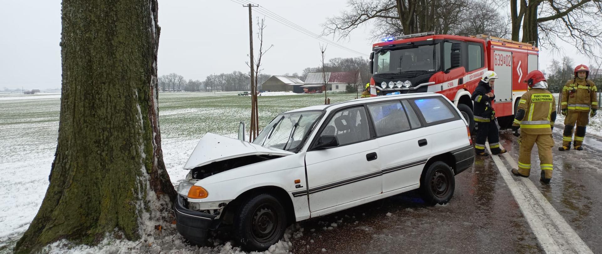 Na zdjęciu widać rozbite auto osobowe na drzewie, strażaków oraz wóz strażacki. 
