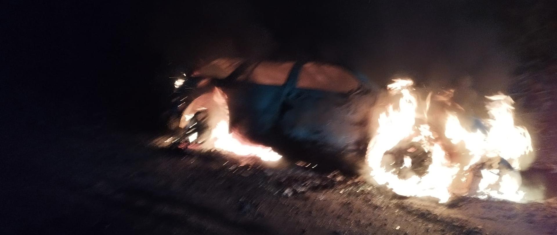 Zdjęcie przedstawia objęty pożarem samochód osobowy w porze nocnej. Samochód znajduje się w rowie.