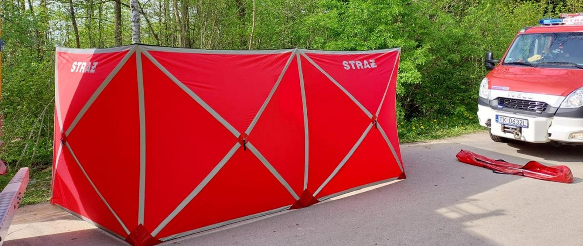 Zdjęcie przedstawia rozstawiony, czerwony parawan, który zasłania miejsce zdarzenia. Z prawej strony stoi zaparkowany samochód straży pożarnej.
