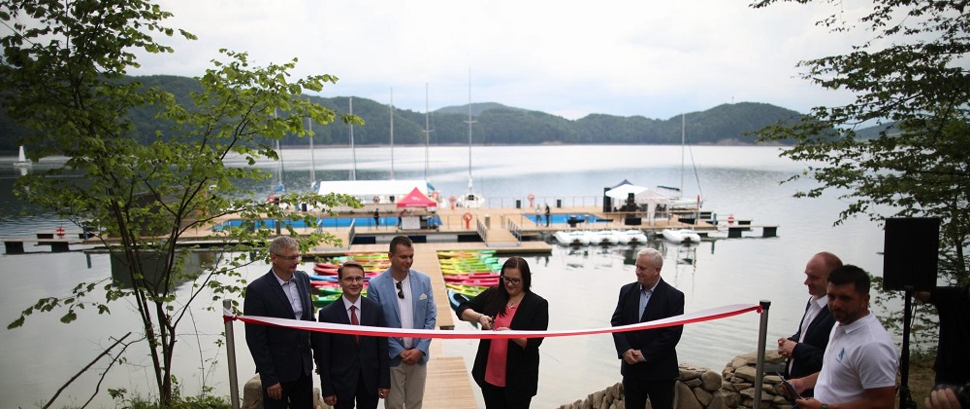 Zdjęcie jeziora i mariny. Biało-czerwoną wstęgę przecina minister Jarosińska-Jedynak, na zdjęciu stoi obok 6 mężczyzn.