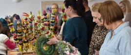 Grupa osób zgromadzonych wokół dużego stołu podczas kiermaszu ozdób wielkanocnych. Na blatach znajdują się liczne dekoracje, głównie kolorowe palmy wykonane z barwionych zbóż, suszonych roślin i kwiatowych elementów. Palmy stoją gęsto w wiklinowej skrzyni, tworząc efektowną, wielobarwną ekspozycję pełną odcieni żółci, różu, zieleni, fioletu, czerwieni i turkusu. Kilka osób ogląda oraz trzyma w dłoniach wiosenne wianki z zielonych gałązek i pastelowych kwiatów. Jedna z osób podnosi zieloną obręcz z dekoracjami, inna trzyma wianek utrzymany w jasnych, delikatnych barwach. Widać jasne przestronne pomieszczenie z dużymi oknami po prawej stronie, które zapewniają naturalne oświetlenie. W tle widoczne są dodatkowe stoły z dekoracjami oraz fragmenty wiszących obrazów. 