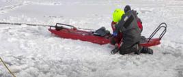 Zdjęcie przedstawia wydobytą z wody przez ratownika osobę, pod którą załamał się lód. Osoba z ratownikiem znajdują się przy saniach lodowych. Wokół przerębla znajdują dwa przęsła drabiny nasadkowej 