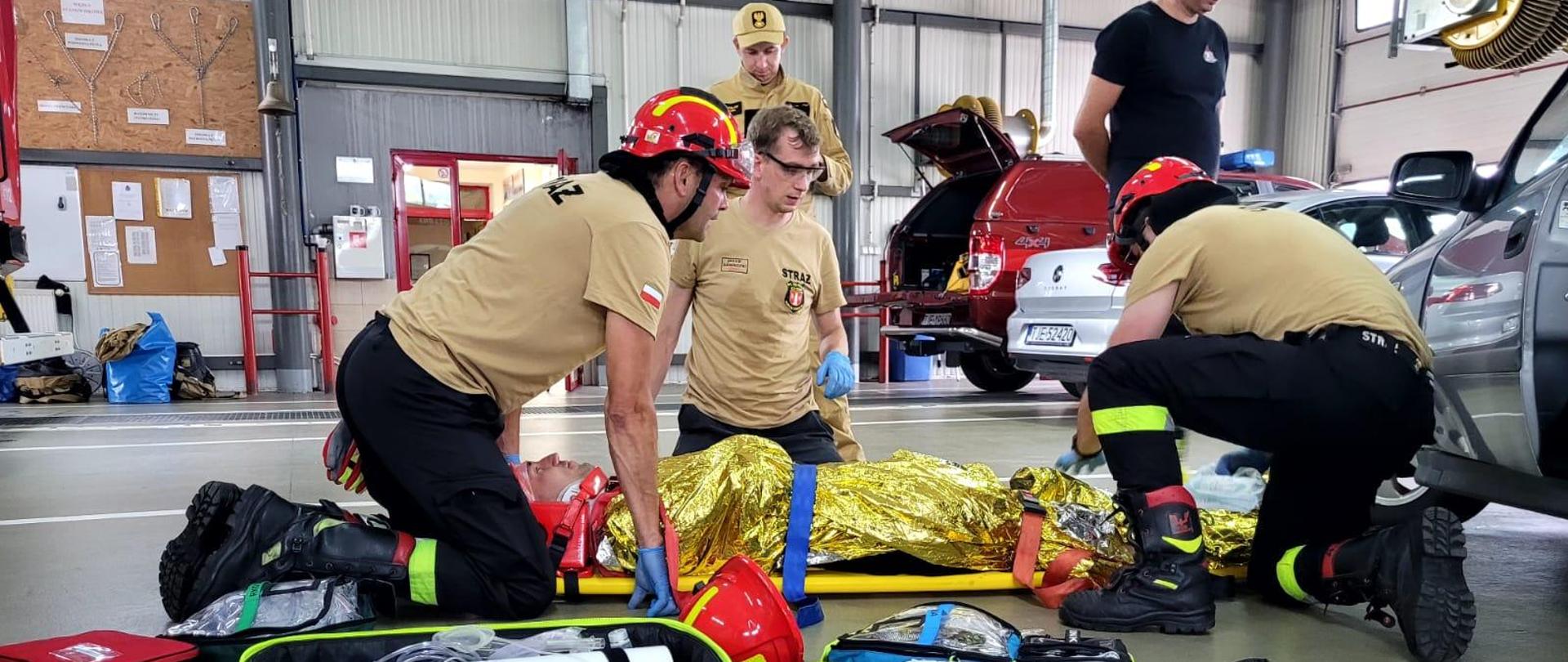 Na desce zaopatrzony medycznie poszkodowany. Przy nim ratownicy w czerwonych hełmach, obok rozłożona torba medyczna R1 oraz fantom noworodka.