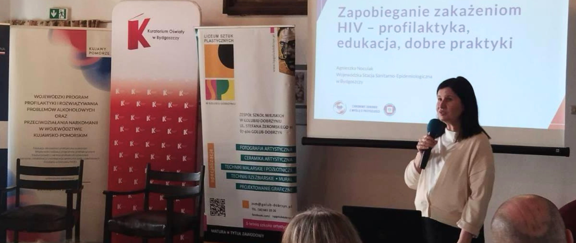 Agnieszka Noculak stojąca przy ekranie projekcyjnym prowadzi prezentację na temat profilaktyki HIV, trzymając mikrofon w ręku. Obok znajdują się roll-upy instytucji edukacyjnych, a na slajdzie widnieje tytuł dotyczący zapobiegania zakażeniom HIV. Przed nią siedzą uczestnicy spotkania.
