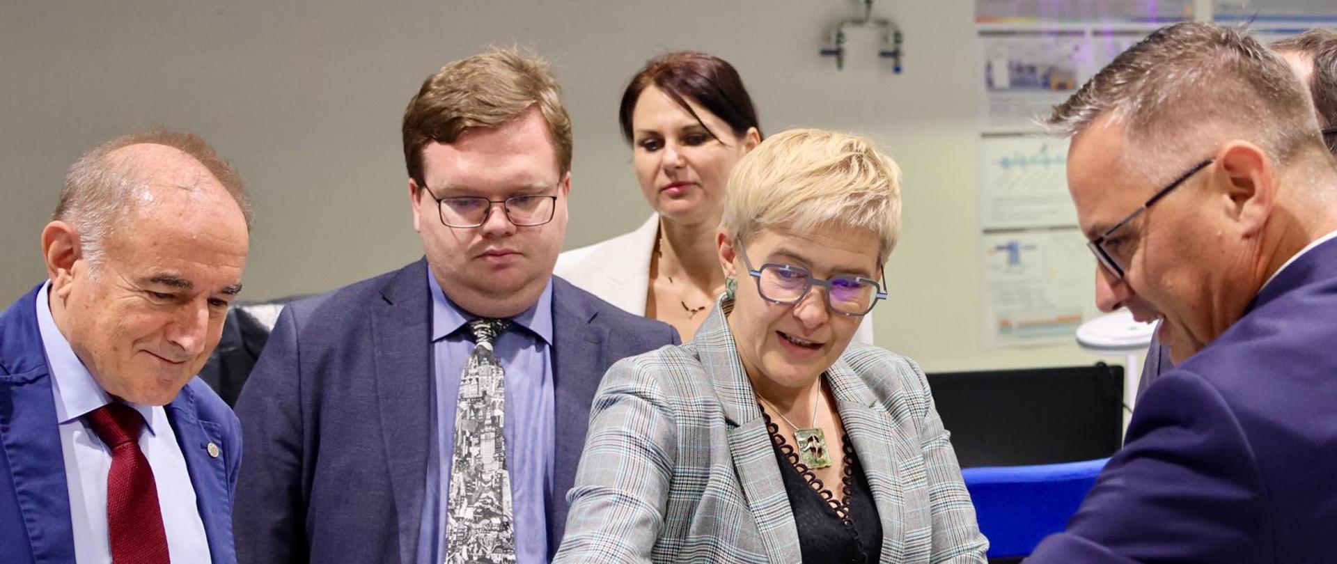 Wiceminister Mrówczyńska i trzech mężczyzn w garniturach stoją i rozmawiają nad otwartym laptopem.