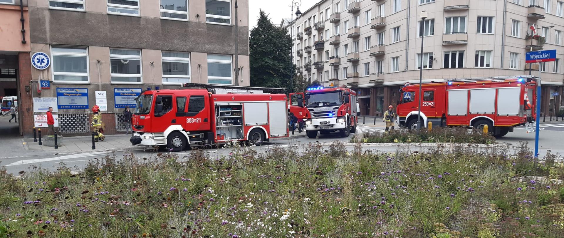 Na zdjęciu na pierwszym planie widać 3 samochody gaśnicze koloru czerwono – białego przed budynkiem Szpitala. Na zdjęciu widać też ratowników w ubraniach specjalnych koloru piaskowego którzy prowadzą działania gaśnicze.