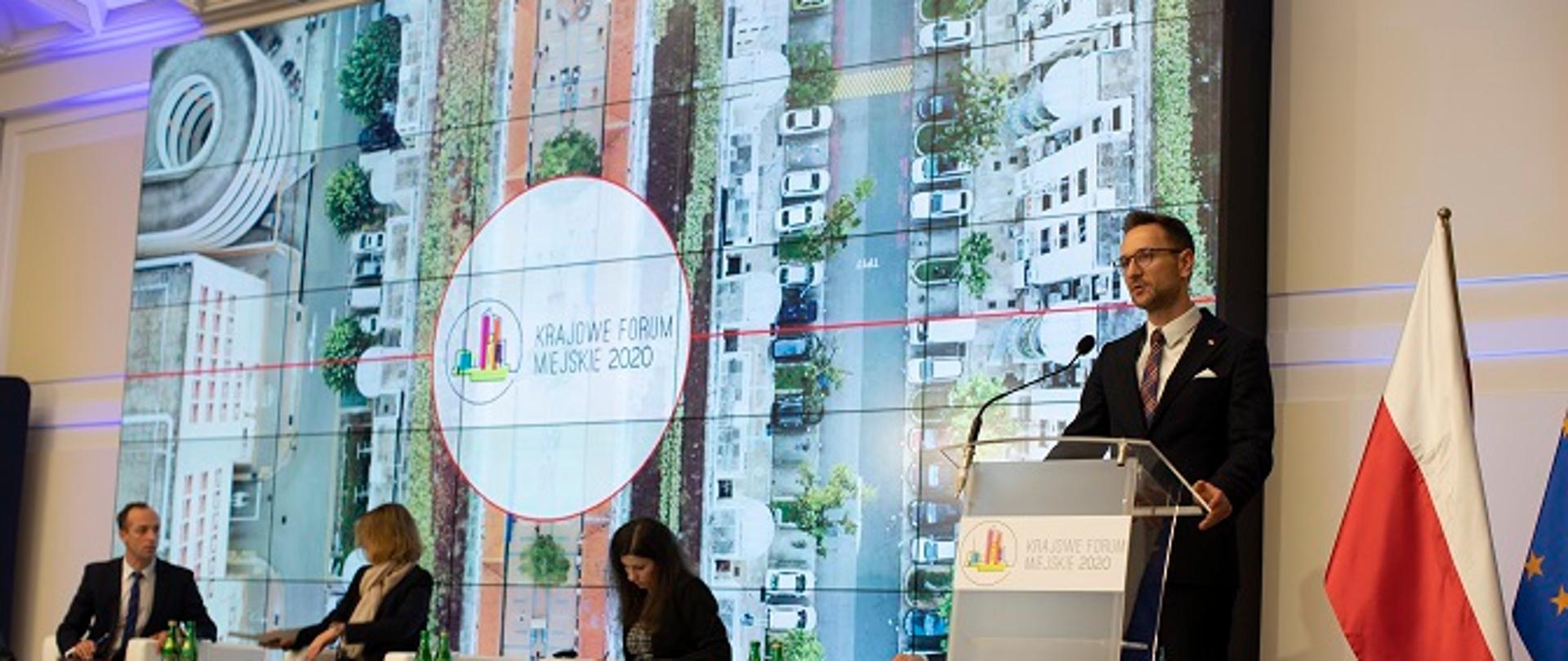 Zdjęcie przedstawia wiceministra Waldemara Budę przemawiającego na Krajowym Forum Miejskim 2020. W tle zdjęcie miasta z góry. Obok dwie kobiety i mężczyzna siedzący na fotelach.