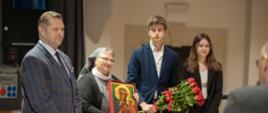 Minister Czarnek w marynarce w kratę stoi obok trzymającej obrazek częstochowskiej Matki Boskiej zakonnicy ubranej na szaro, za nimi dwoje młodych ludzi, mężczyzna trzyma bukiet czerwonych kwiatów.