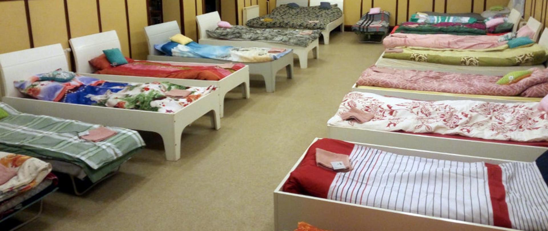 Na zdjęciu widoczna jest sala w budynku ochotniczej straży pożarnej. Po obydwu stronach, w rzędach ustawione są łóżka, na nich leżą złożone ręczniki i pościel. Na ścianach wiszą zdjęcia i dyplomy.