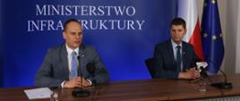 Na zdjęciu widać dwóch mężczyzn, którzy siedzą przy stole. Przed nimi stoją mikrofony, a z tyłu ścianka z napisem Ministerstwo Infrastruktury. Po lewej stronie, widać biało-czerwoną flagę oraz flagę Unii Europejskiej