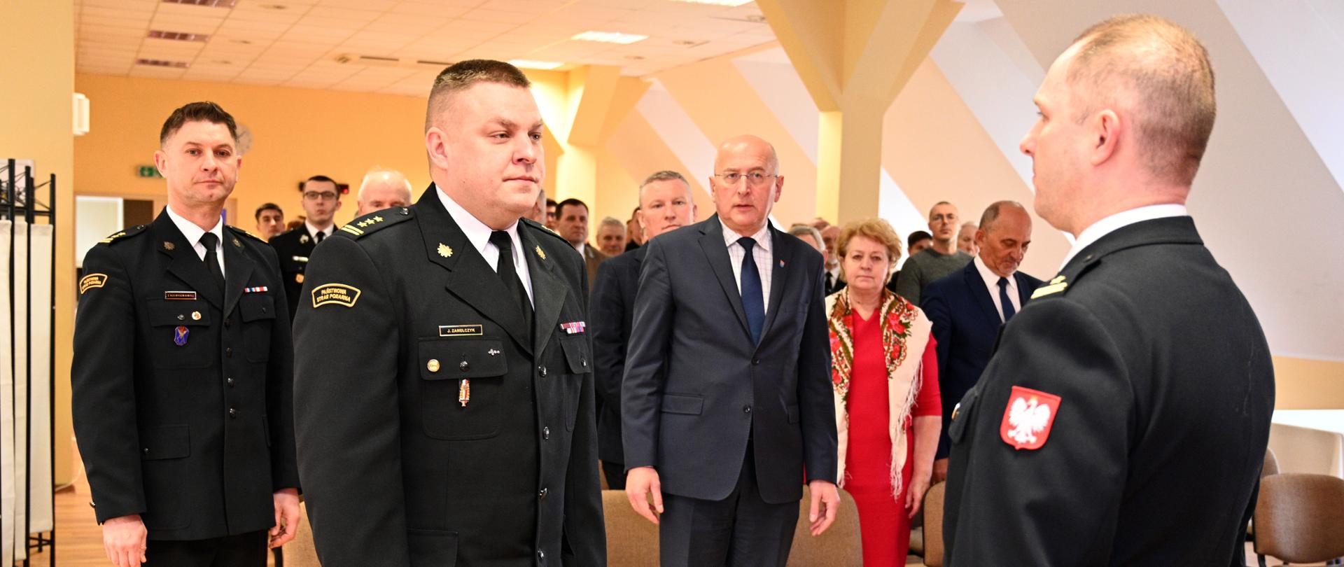 w sali, składanie meldunku strażakowi przez drugiego strażaka, za nimi grupa ludzi