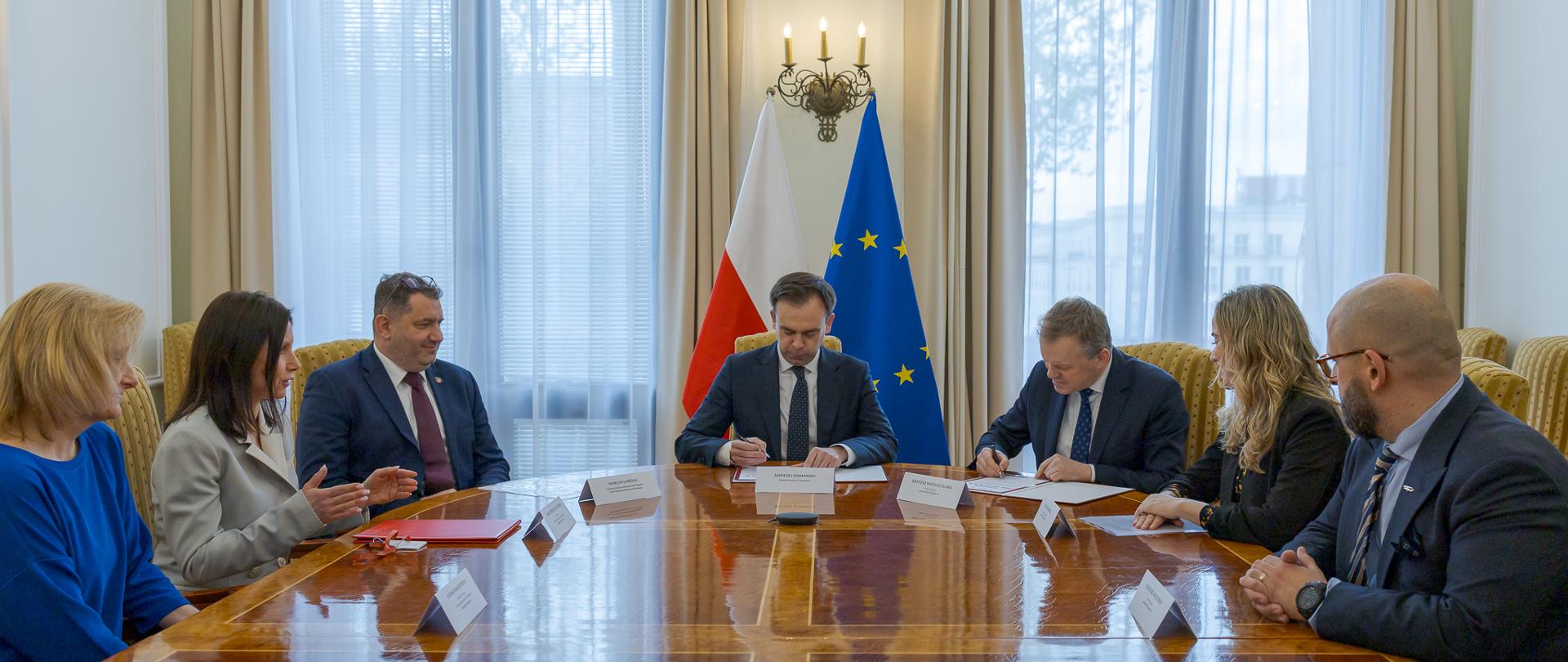 Uczestnicy spotkania w tym Minister Finansów i Gospodarki Andrzej Domański siedzą przy stole konferencyjnym w sali z flagą Polski i Unii Europejskiej.