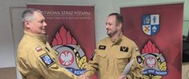 Pomieszczenie świetlicy w komendzie powiatowej państwowej straży pożarnej w sępólnie krajeńskim, Komendant Powiatowy gratuluje wyróżnionemu funkcjonariuszowi który trzyma w ręku wyciąg z rozkazu służbowego. 