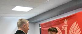 Strażak w mundurze wyjściowym gratuluje chłopakowi i wręcza dyplom. W prawej części zdjęcia czerwony baner PSP. W tle przed białą ścianą stoją inni uczestnicy turnieju. 