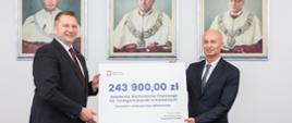 Minister Czarnek i mężczyzna w garniturze stoją i trzymają razem symboliczny czek z napisem 243 900 zł, za nimi na ścianie wiszą 3 portrety.