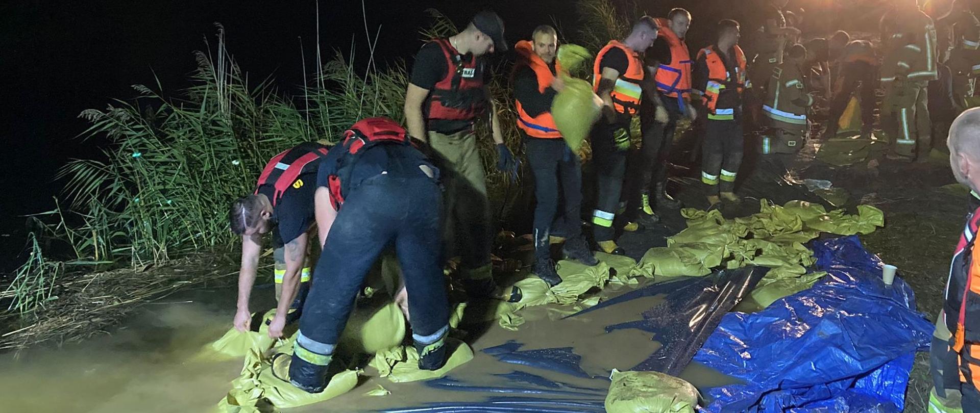 Strażacy układają worki z piaskiem na koronie wału, częściowo stoją w wodzie. Teren akcji oświetlają strażackie najaśnice.
