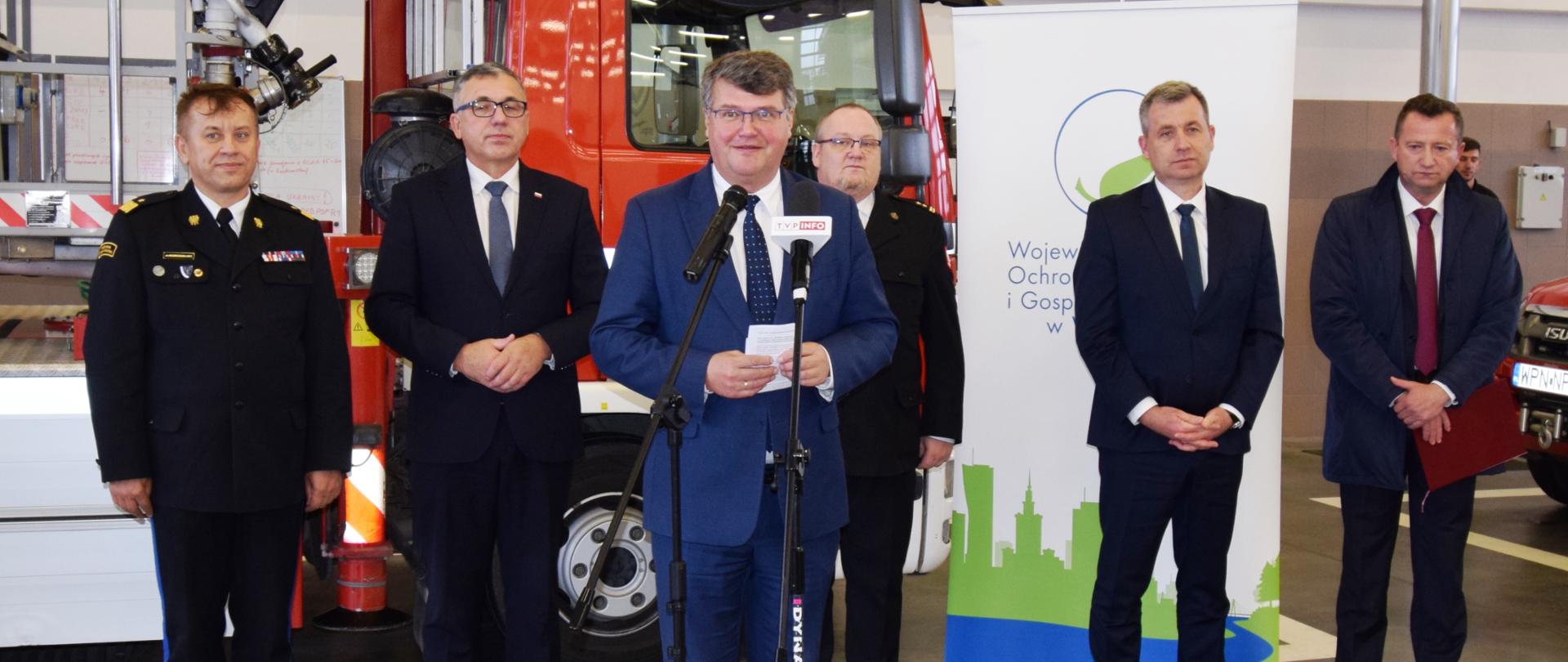 Jedna osoba w niebieskim garniturze przemawia przed mikrofonem. Z tyłu stoi pięć osób w ciemnych garniturach. W tle czerwony samochód pożarniczy. 