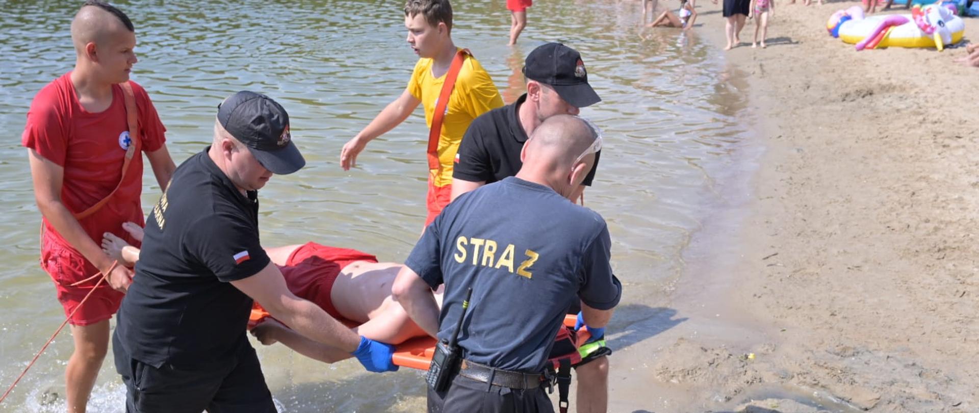 Na zdjęciu widoczni strażacy i ratownicy wodni wynoszący na desce z wody osobę poszkodowaną.