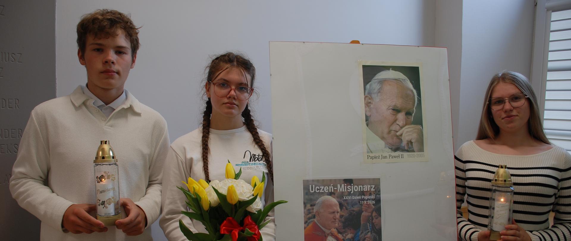 Uczeń i dwie uczennice ze zniczem i kwiatami przed plakatem o Dniu Papieskim 