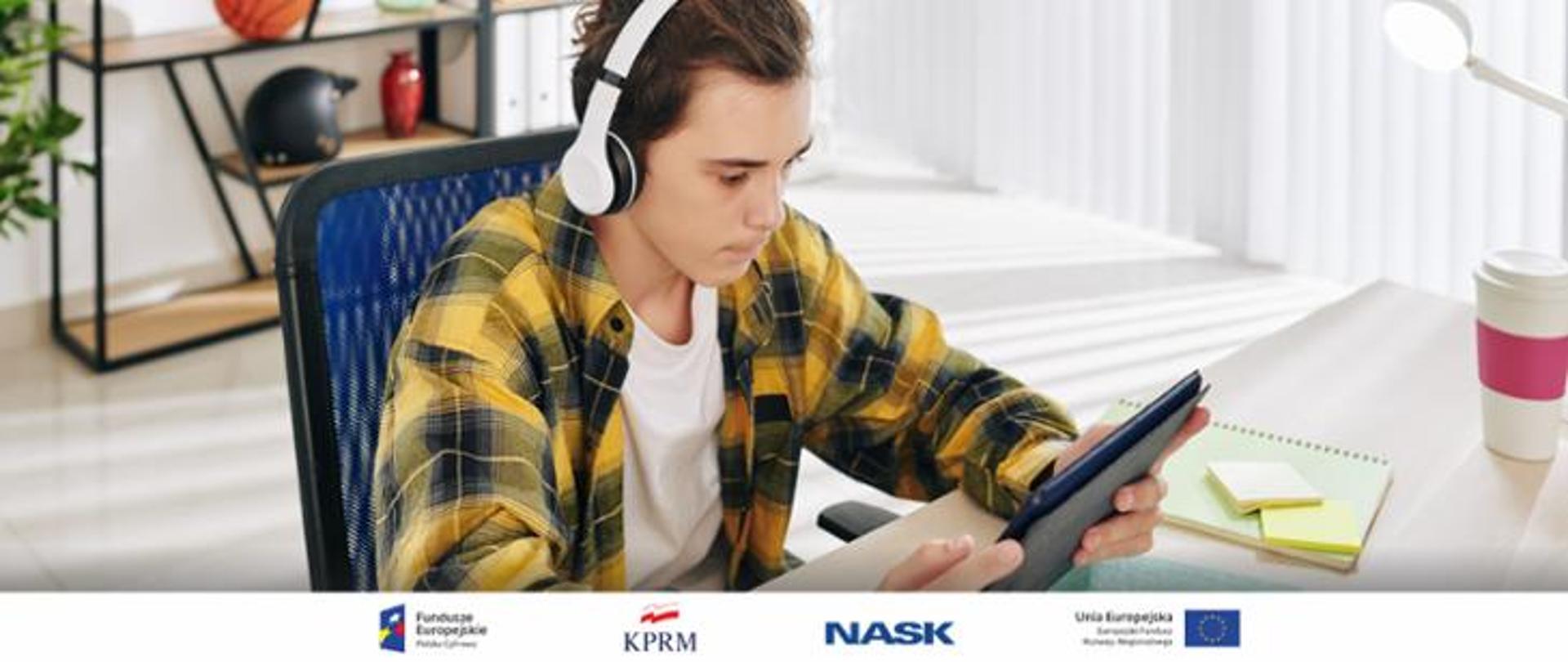 
Zdjęcie nastolatka w słuchawkach siedzącego przy biurku - w rękach trzyma tablet, na którym coś ogląda