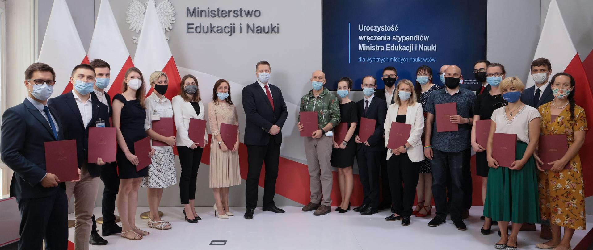 Minister Czarnek stoi w otoczeniu młodych naukowców, za nimi na ścianie napis Ministerstwo Edukacji i Nauki i polskie flagi.