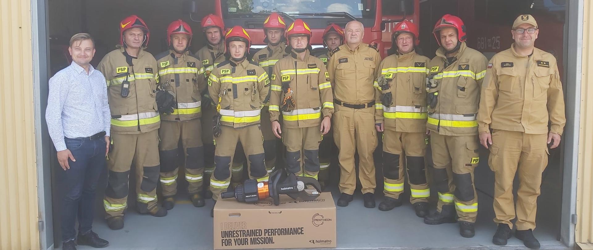 Zdjęcie przedstawia zmianę służbową strażaków JRG Wołomin w obecności Komendanta Powiatowego PSP w Wołominie oraz przedstawiciela firmy Inter Europol S.A. Strażacy otrzymują narzędzie hydrauliczne. Zdjęcie wykonane na tle pojazdu strażackiego stojącego w garażu jrg.