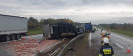 Przewrócona cysterna leży na pasie drogi i barierkach, po lewej widać naczepę, z przodu stoi ciągnik siodłowy, po prawej widać czterech strażaków