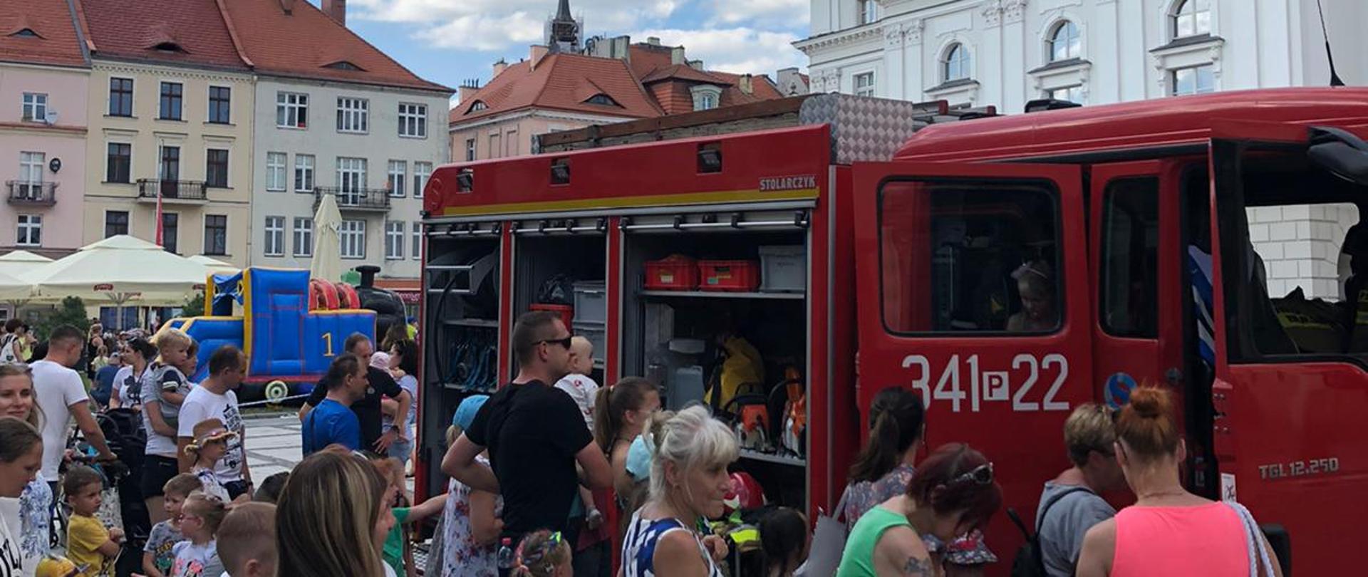 Na pierwszym planie grupa dzieci, które oczekują na wejście do wozu strażackiego. Za kolejką widać samochód strażacki, który posiada kolor czerwony, oznaczenia numerów operacyjnych i sygnalizację ostrzegawczą. W dalszej perspektywie budynek kaliskiego ratusza, który jest w kolorze szarym. Widać zegar i wieżę ratuszową. W oddali dmuchany zamek.
