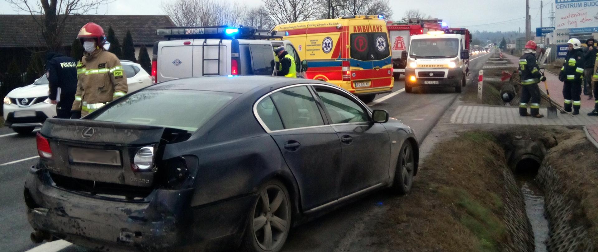 Na pierwszym planie znajduje się samochód osobowy marki Lexus z uszkodzonym tylnym zderzakiem. W tle znajduje się strażak oraz samochody Policji, Pogotowia Ratunkowego i Straży Pożarnej. Po prawej stronie znajdują się strażacy porządkujący teren akcji.