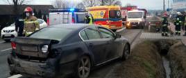 Na pierwszym planie znajduje się samochód osobowy marki Lexus z uszkodzonym tylnym zderzakiem. W tle znajduje się strażak oraz samochody Policji, Pogotowia Ratunkowego i Straży Pożarnej. Po prawej stronie znajdują się strażacy porządkujący teren akcji.