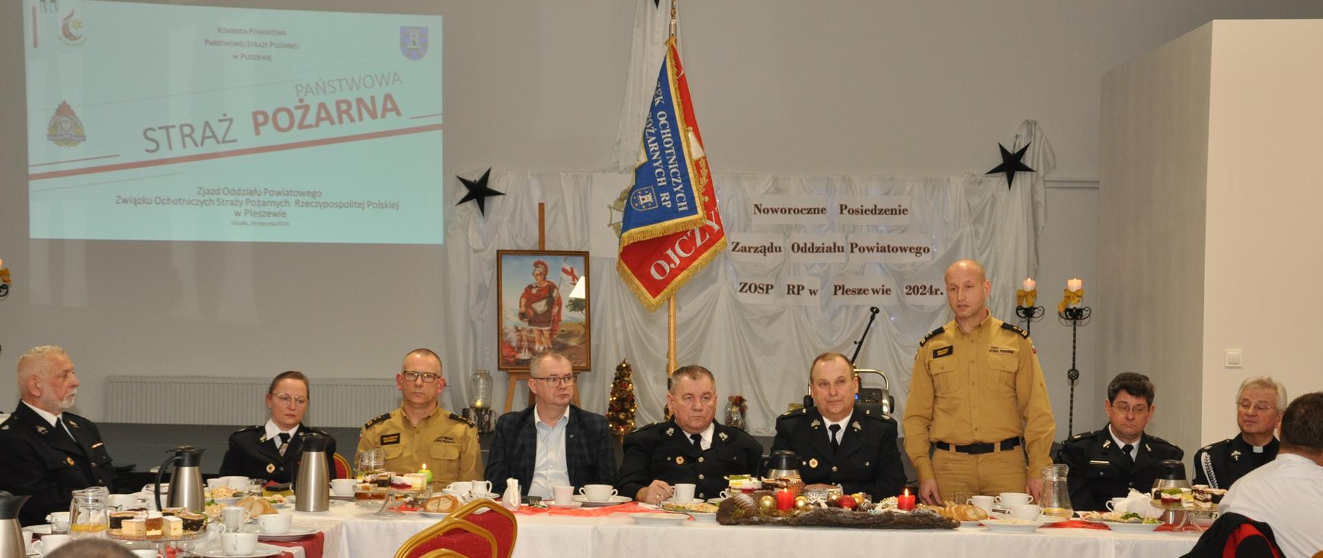 Grupa mężczyzn i kobiet siedzi w oświetlonej sali za stołem prezydialnym, jeden mężczyzna ubrany w mundur państwowej straży pożarnej przemawia na stojąco