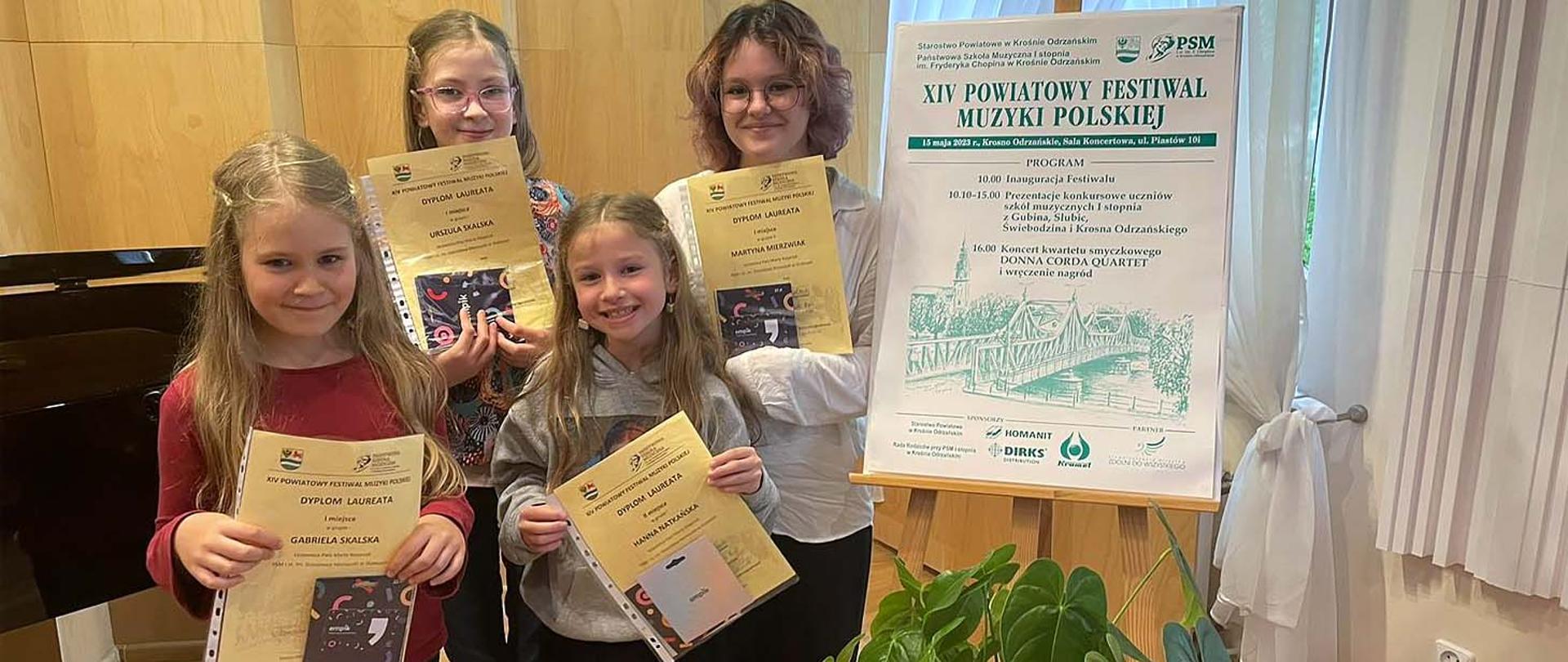 Pięć dziewczynek stoi obok plakatu reklamującego Festiwal Muzyki Polskiej, wszystkie z nich w rękach trzymają dyplomy.